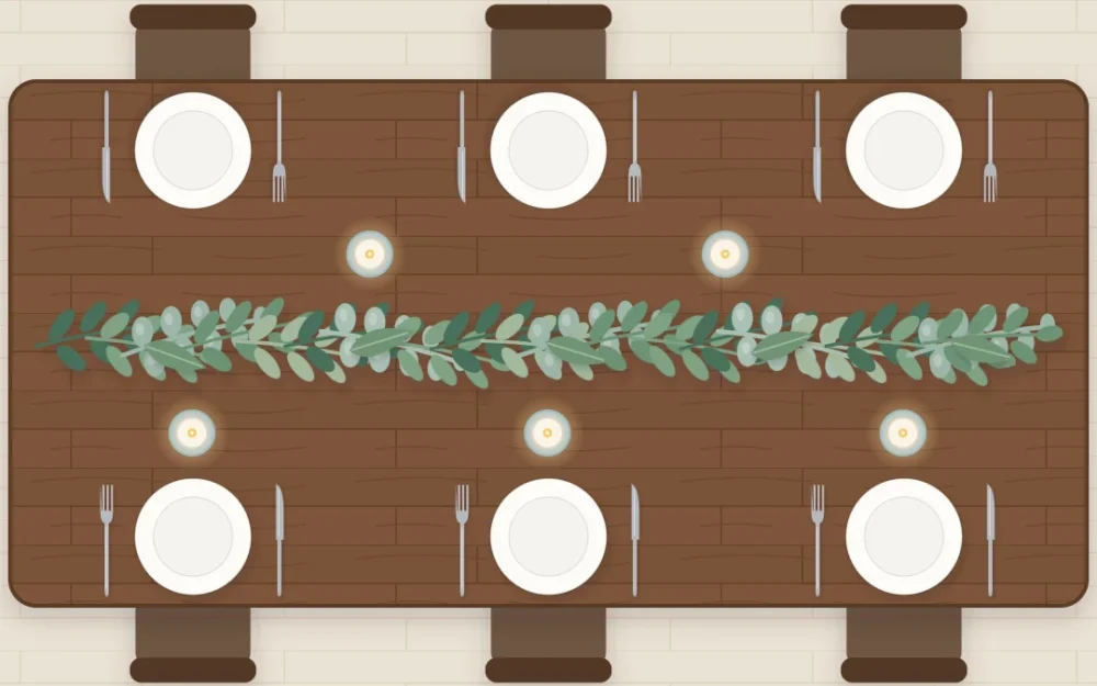 Top-down view of a long wooden table with a loose garland of mixed green leaves down the center, small flameless candles in glass holders and ivory plates